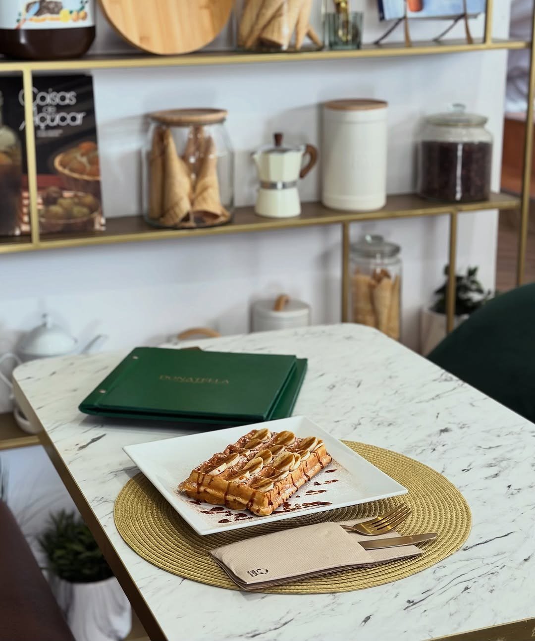 Mesa de mármore com um waffle e a carta verde da casa, com prateleiras de frascos ao fundo